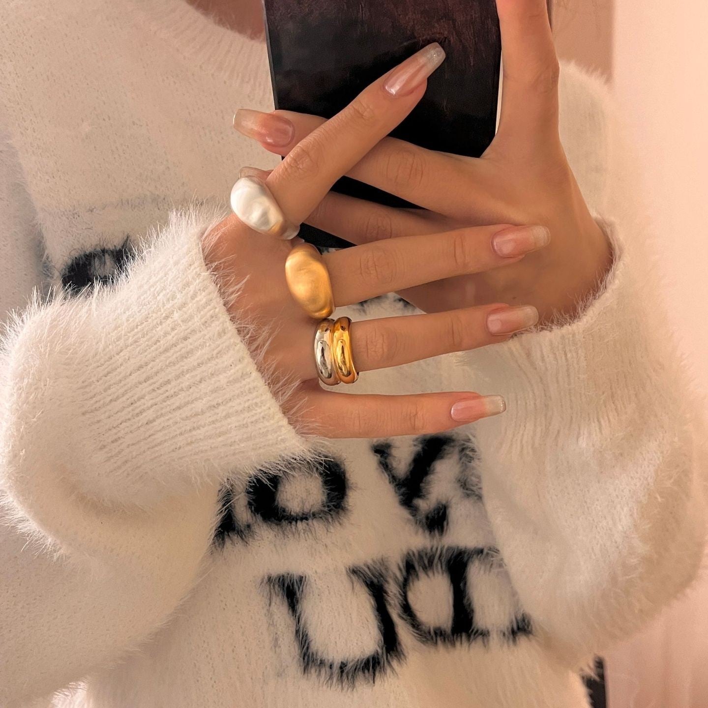 Close-up of a hand with rings holding a phone, wearing a white sweater with black text.