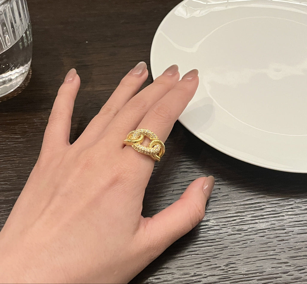 Hand wearing a gold ring on a wooden surface with a white plate and glass in the background.