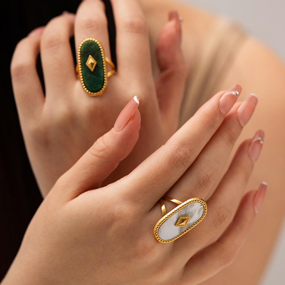 Close-up of hands wearing gold rings with green and white stones on a neutral background